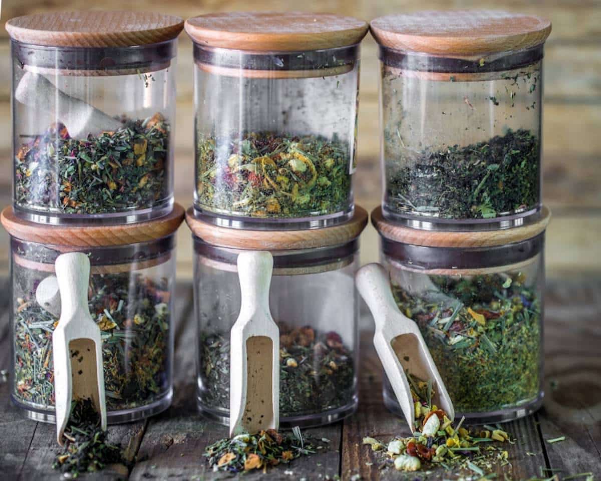 Four glass jars with wooden lids, containing various dried herbs and teas, each with a wooden scoop.