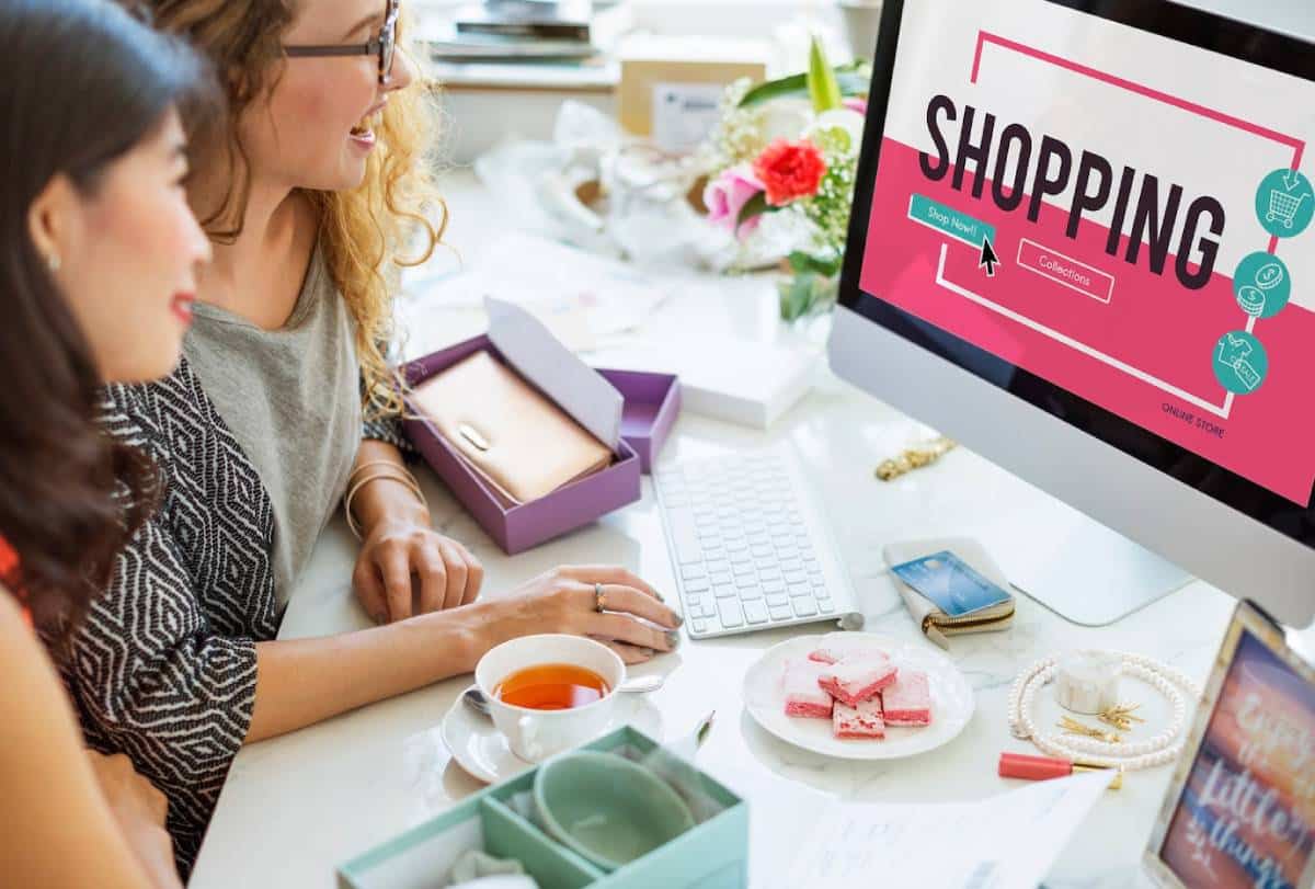 Two women shopping online on a computer, with jewellery and snacks on the table.