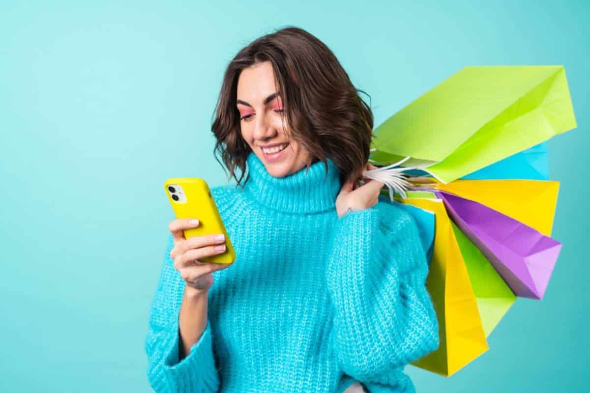 Woman in blue jumper smiles at phone, holding colourful shopping bags, against turquoise background.