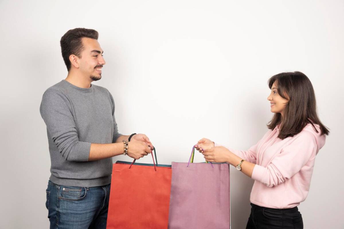 Two people, a man and a woman, smiling and holding colorful shopping bags, against a grey background. 