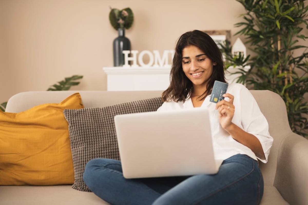 Woman sits on a sofa, using a laptop and holding a credit card, smiling in a cozy home setting.