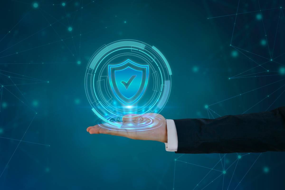A person's hand holding a holographic shield symbolizing cybersecurity, set against a digital blue background.