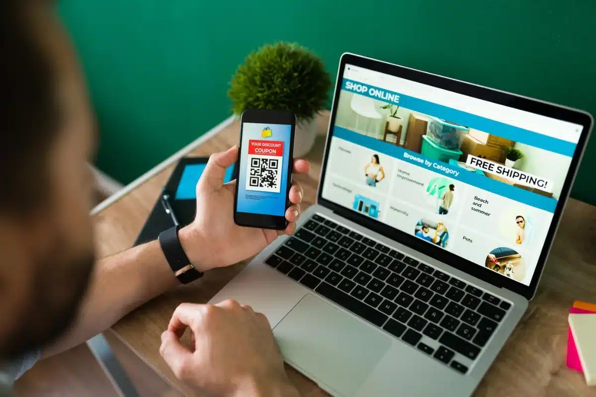 Man using smartphone to scan QR code for discounts while browsing online shopping site on laptop.