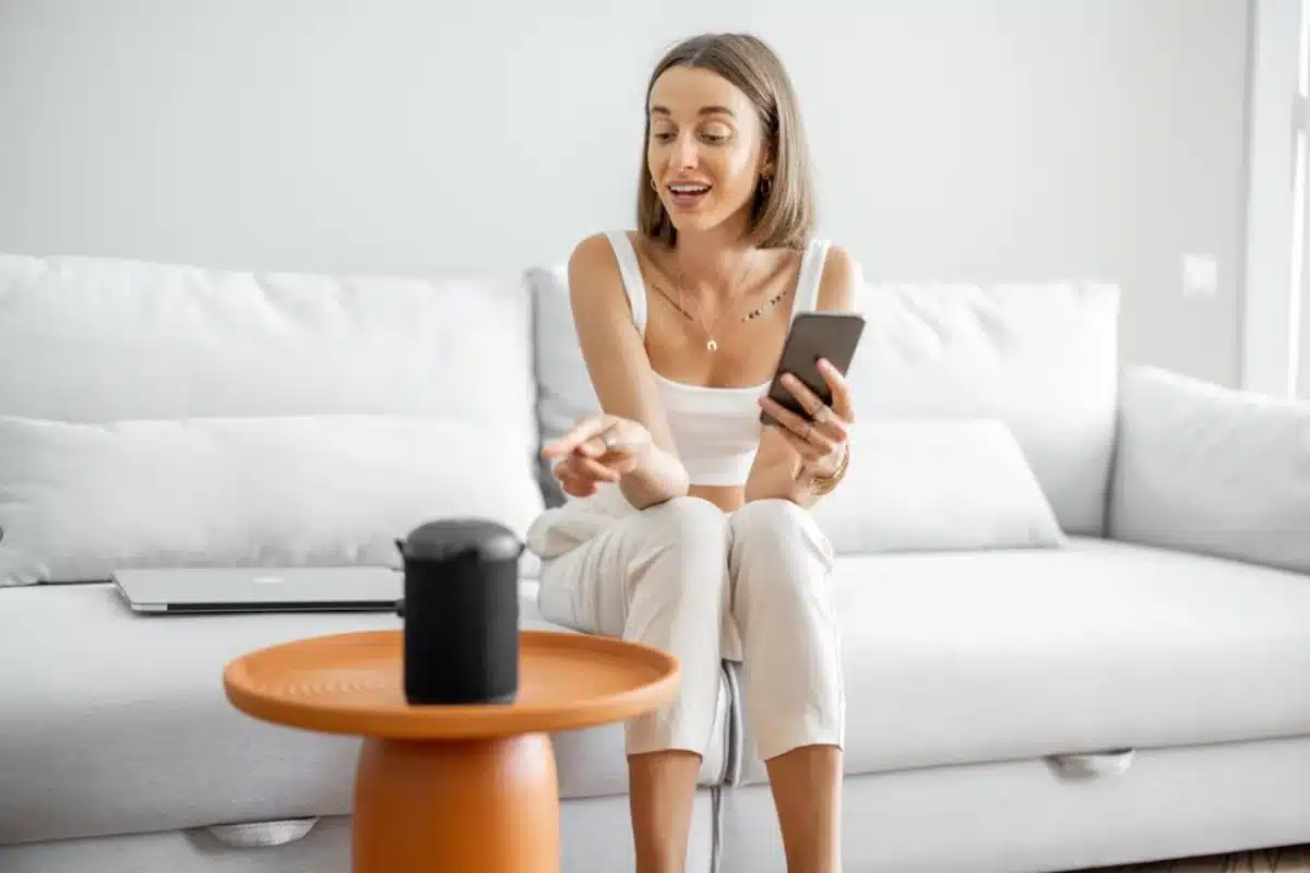 Woman sitting on a sofa, speaking into a smart speaker while holding a smartphone, in a modern living room.