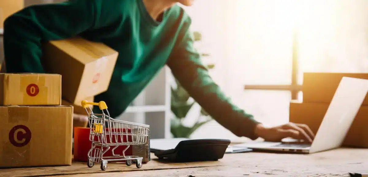 Person in a green sweater packing boxes and using laptop in a sunny workspace, with a miniature shopping trolley beside them.