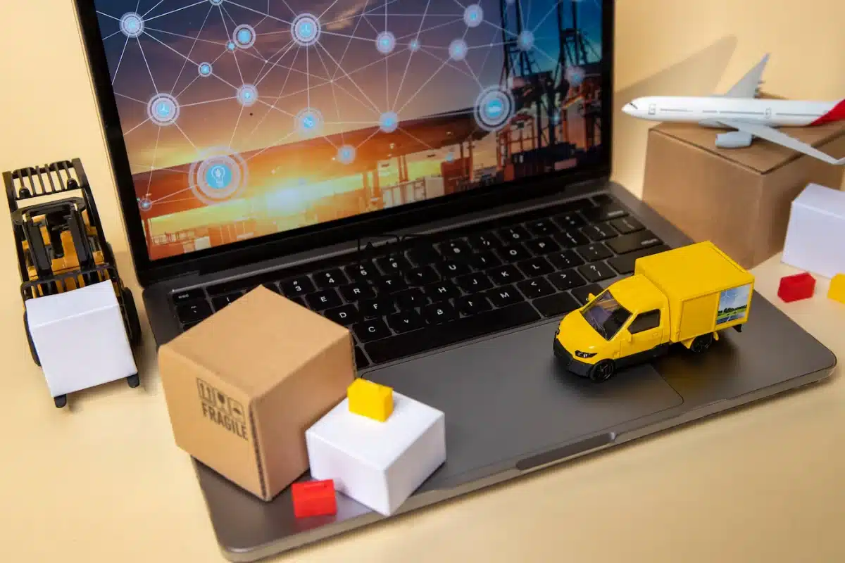 Model logistics setup with a laptop screen displaying a map, plane model, yellow delivery van, and various small packages on a desk.